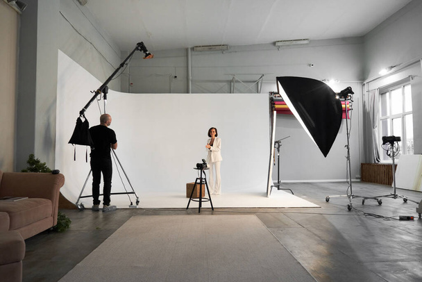 Fashion photography in a photo studio. Professional male photographer taking pictures of beautiful woman model on camera, backstage - Photo, Image