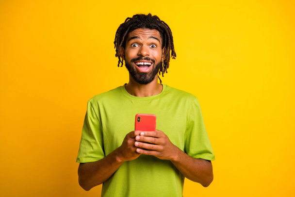 Photo of astonished dark skin guy hold smartphone impressed wear green t-shirt isolated on shine yellow color background - Foto, afbeelding