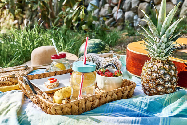 Summertime healthy picnic. Fresh tropical fruit, sweet stawberry sandwiches and refreshing drink in mason jar on wicker tray on blue cloth in the garden. - Photo, Image