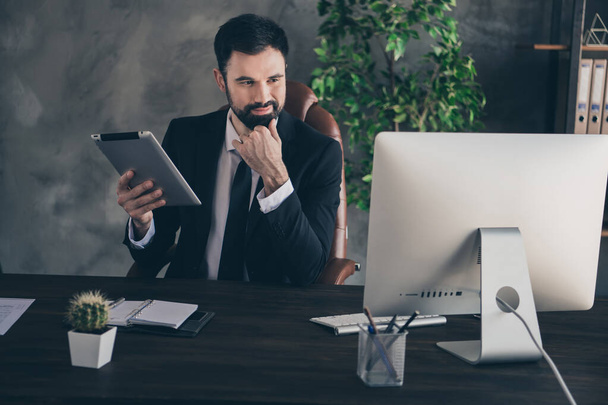 Foto de hombre exitoso sentarse escritorio mantenga tableta dedo barbilla mirada pc pantalla desgaste traje camisa corbata en la oficina moderna en interiores - Foto, Imagen