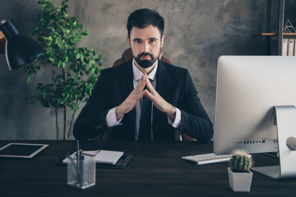 Zdjęcie pewny siebie agent człowiek siedzieć biurko trzymać palce chłodny wygląd aparat nosić garnitur koszula krawat w nowoczesnym biurze w pomieszczeniach - Zdjęcie, obraz