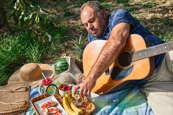 Pareja de mediana edad haciendo un picnic en el jardín con fruta exótica fresca y sándwiches dulces. Hombre en camiseta azul con patrón de flores toca la guitarra. Primavera, verano estilo de vida, amor citas - Foto, Imagen