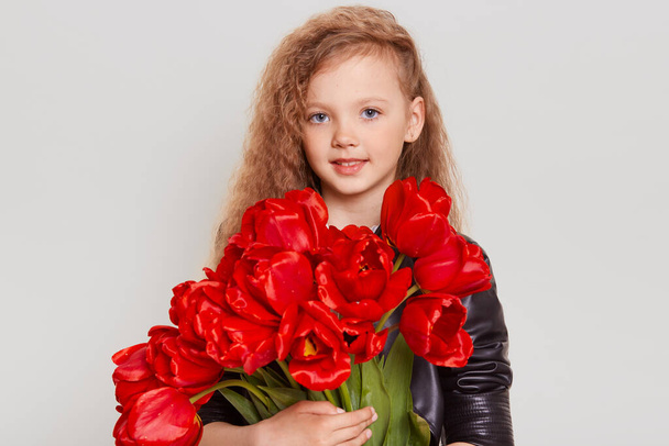 Pleasant looking blonde wavy haired child in black leather jacket, looking at camera with smile, embracing red tulips in hands, wants to give it for somebody, isolated over gray background. - Photo, image