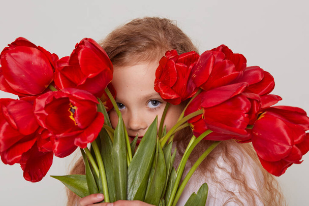 Beautiful blonde girl with wavy hair having fun alone, hiding behind big bouquet of tulips, looking at camera as a spy, isolated over gray background. - Fotó, kép