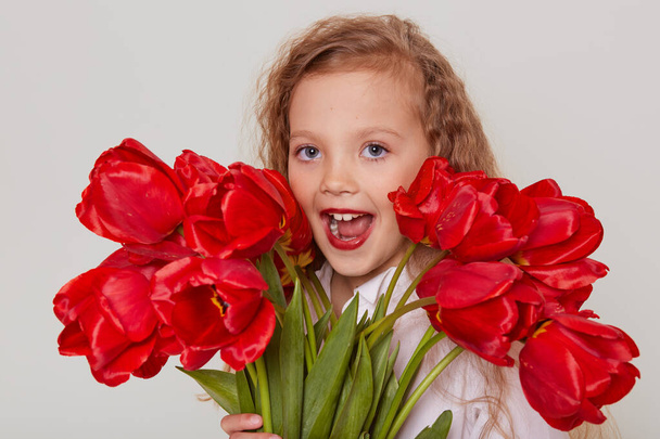 Excited schoolgirl with red tulips in hands, looking at camera with widely opened mouth, having fun alone, being happy to get such present, isolated over gray background. - Foto, imagen