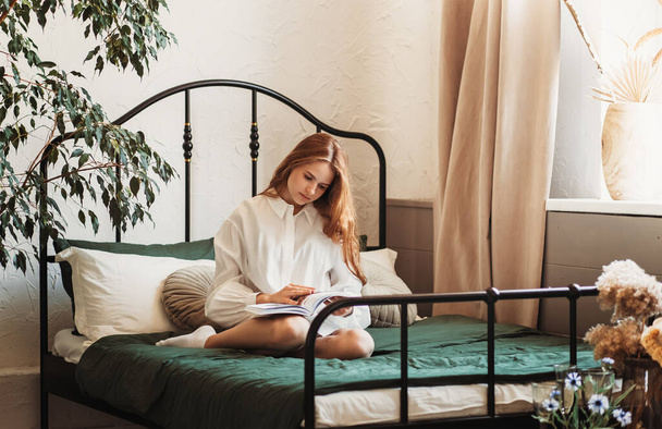 Uma jovem romântica com cabelos longos se senta em uma cama com um livro. Ler na cama - Foto, Imagem