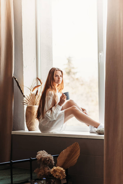 Een jong meisje met lang haar zit op een vensterbank met een beker in haar handen - Foto, afbeelding