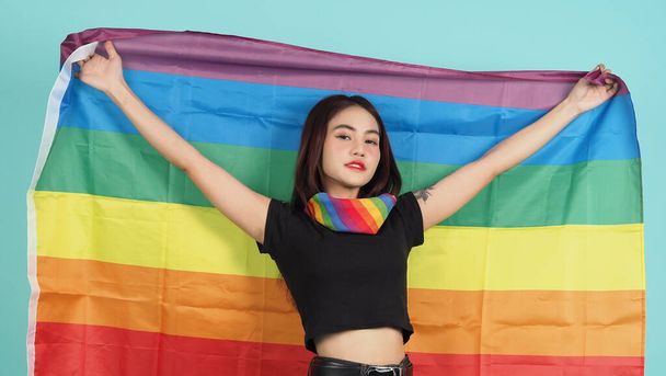 Chica LGBTQ y bandera de orgullo. Sexy chica lesbiana y bandera LGBT de pie. fondo verde azul. Mujer LGBTQ asiática con bufanda arcoíris en el cuello. Enérgico alegre. Concepto LGBTQ.  - Foto, imagen
