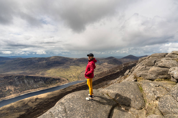 Mujer sonriente con gorra, chaqueta rosa, polainas amarillas de pie en la cima de la montaña, con lago en el fondo en Slieve Binnian Mountain Irlanda del Norte - Foto, imagen