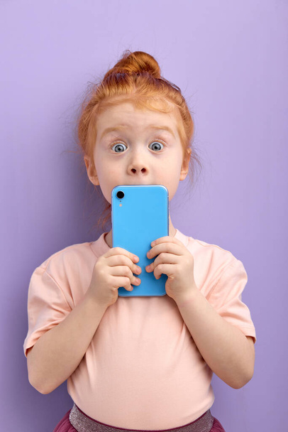 Portrait d'enfant rousse surprise utilisant un smartphone, isolé sur fond violet en studio. - Photo, image