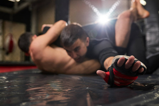 Young MMA fighters grappling on training in gym. Muscular sportsman lying in floor and resisting, close-up hands - Photo, image