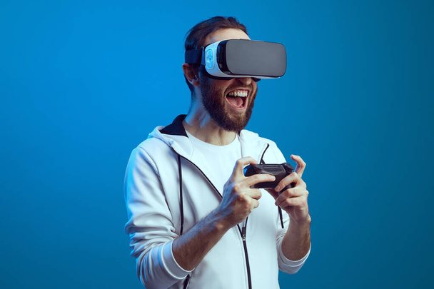 Joven hombre barbudo emocionado de pie con el equipo VR y jugando - Foto, imagen