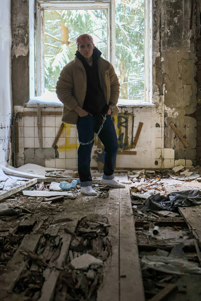 The young man with pink blonde hair in fluffy coat and black hoodie standing near the window without glass in old ruin abandoned urban building - Φωτογραφία, εικόνα