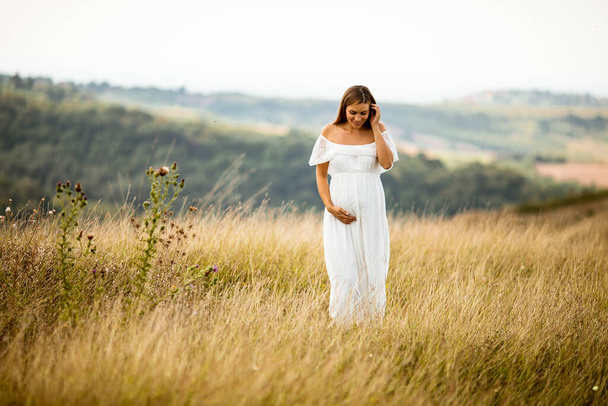 Молода вагітна жінка в білій сукні на літньому полі
 - Фото, зображення