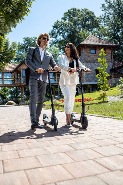 Young fancy dressed couple having fun while pushing their electric scooters, businesspeople on the road concept  - Foto, Imagem