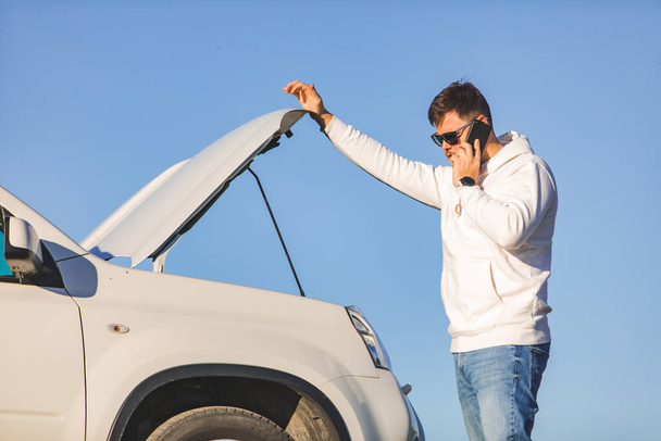 Мужчина, стоящий рядом с машиной с открытым капюшоном на закате, разговаривает по телефону - Фото, изображение