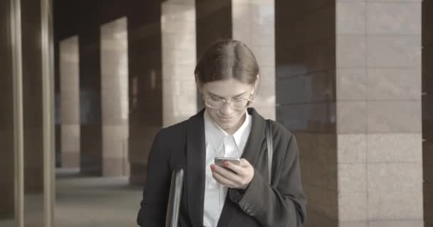 Relaxed successful businesswoman in suit and eyeglasses browsing and texting on the mobile phone while walking toward the camera. 4k. - Séquence, vidéo