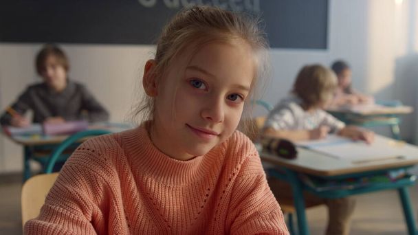 Aranyos diák ül az általános iskola íróasztalánál. Mosolygó lány a kamerába néz  - Fotó, kép