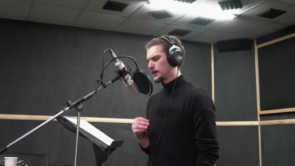 Curly joven en auriculares canta cerca del micrófono en el estudio de grabación de voz  - Imágenes, Vídeo