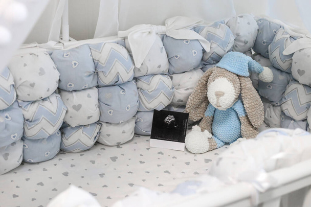 Leeres Babybett mit schönen blauen Textilien. Ultraschall eines Kindes mit Strickspielzeug. Schwangerschaft. Baldachin und weiße Krippe. - Foto, Bild
