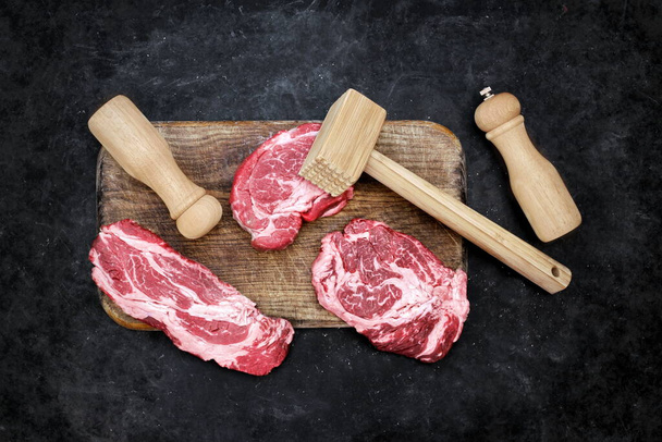 Μπριζόλες βοδινού, Ξύλινο Σφυρί, Αλάτι και Πιπέρι στο ξύλο κοπής. Μπριζόλες βοείου κρέατος BBQ ή ψησίματος, Overhead View. Ακατέργαστο Striploin Marbled Beef Steaks σε μαύρο φόντο, πάνω όψη. - Φωτογραφία, εικόνα