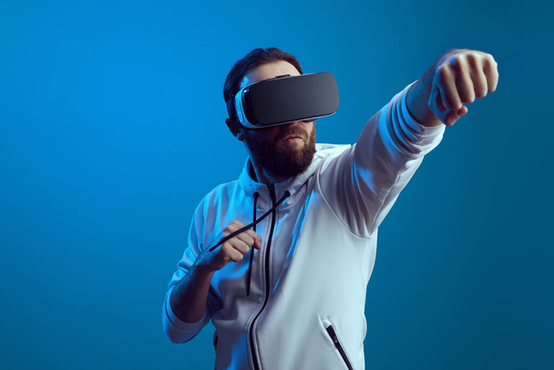 Mladý muž v moderním VR zařízení stojí v boxerské poloze přes modrou stěnu - Fotografie, Obrázek