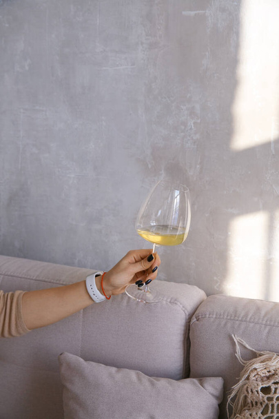 Glass goblet with white wine in a woman's hand against the background of a gray wall near the window - Foto, Bild