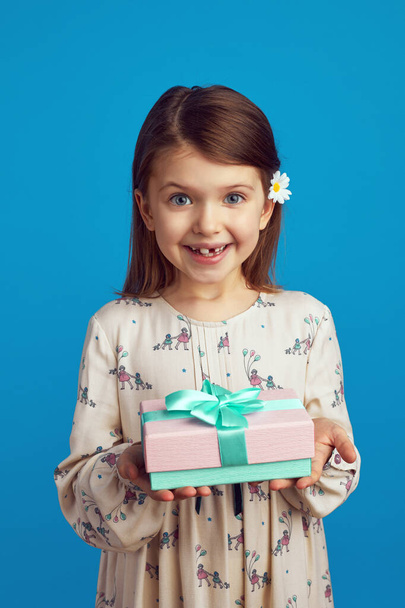 Little girl demonstrating wrapped gift box with ribbon against blue wall - 写真・画像