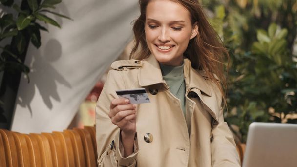 Krásná usměvavá dívka oblečená v trench kabát drží kreditní kartu a usmívá se pracuje na notebooku v kavárně venku. Moderní technologie - Fotografie, Obrázek