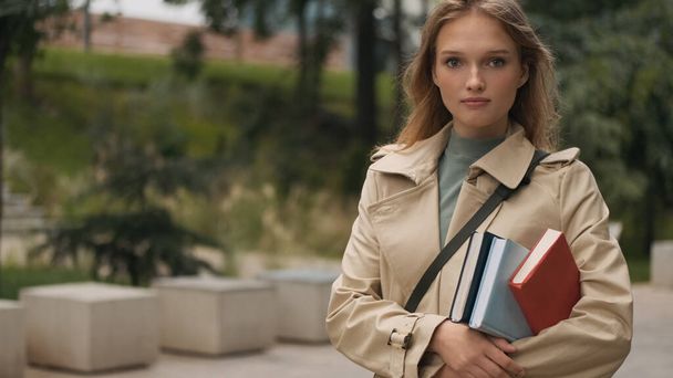 Atraktivní stylová studentská dívka stojící s učebnicemi hledá jistotu v kameře jít na vysokou školu přes městský park - Fotografie, Obrázek