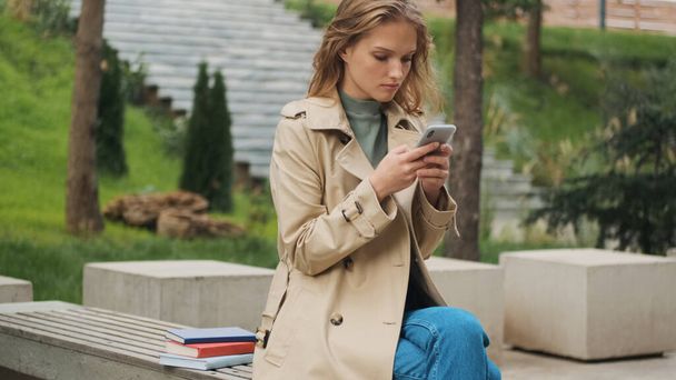 Beautiful stylish student girl chatting with friend on phone sitting on bench with books in park. Study outdoor concept - Photo, Image
