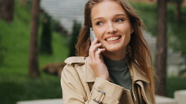 Jeune femme positive regardant élégant parler au téléphone heureux d'entendre le meilleur ami en plein air. Étudiante joyeuse se reposant dans le parc après les cours à l'université - Photo, image
