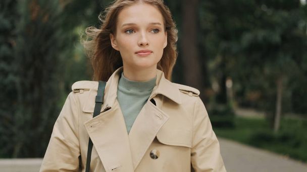 Krásná zamyšlená studentka oblečená v trenč kabát jít na univerzitu přes městský park a oblékání o něčem - Fotografie, Obrázek