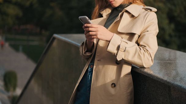 Close up girl dressed in trench coat looking stylish chatting in smartphone outdoor. Sunny weather - Foto, imagen