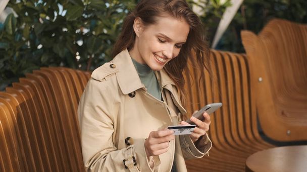 Hermosa chica sonriente mirando feliz pagando con tarjeta de crédito para las compras en línea utilizando el teléfono inteligente en la cafetería de la calle. Concepto de banca por Internet - Foto, imagen
