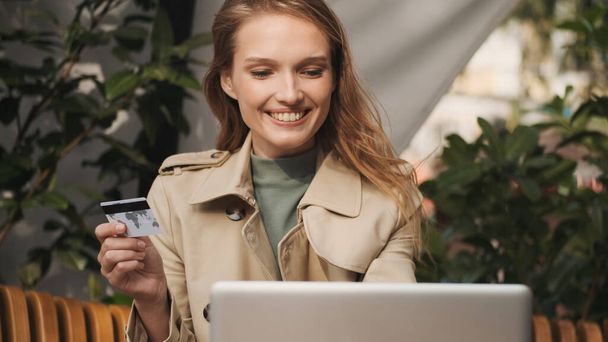 Jovem mulher alegre com cartão de crédito verificando sua conta bancária no laptop usando conexão de internet gratuita no café de rua. Conceito de tecnologia moderna - Foto, Imagem