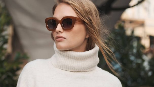 Gorgeous blond girl in sunglasses dressed in white cozy sweater looking confident posing on camera on city street. Stylish lady - Photo, Image