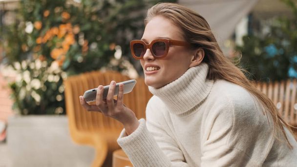 Beautiful stylish girl in sunglasses dressed in cozy white sweater sending voice message to best friend using smartphone in street cafe. Modern technology concept - Valokuva, kuva