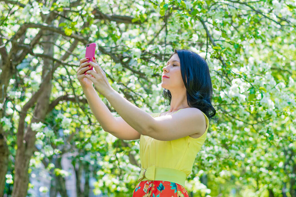 Eine junge Frau in gelbem Kleid steht inmitten einer blühenden Apfelplantage und fotografiert sich mit einem Handy - Foto, Bild