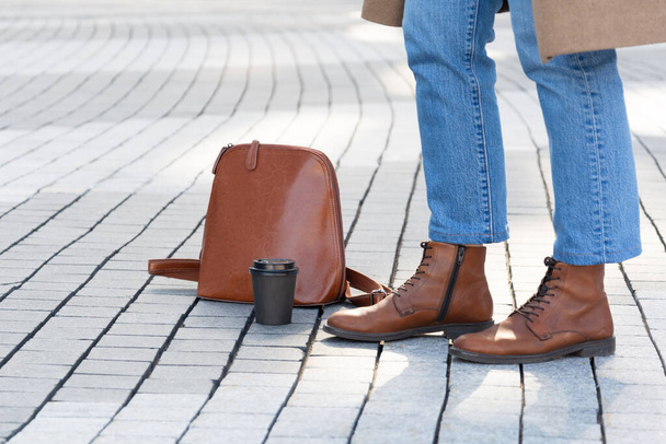 A woman in fashionable clothes with a backpack and a glass of coffee stands on a pavement lined with paving stones - Photo, Image