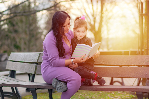 Красивая маленькая девочка читает книгу с матерью и улыбается. Летний размытый парк на заднем плане. - Фото, изображение