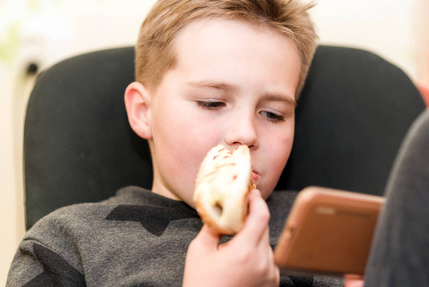 Ένα πεινασμένο αγόρι που τρώει χοτ ντογκ στο σπίτι κάθεται παίζοντας με το smartphone. Εσωτερικό πλάνο. Κλείσιμο.. - Φωτογραφία, εικόνα