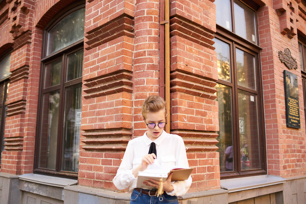 estudiante femenina con gafas de pelo corto cerca de edificio de ladrillo en la calle y camisa blanca corbata vista recortada - Foto, Imagen