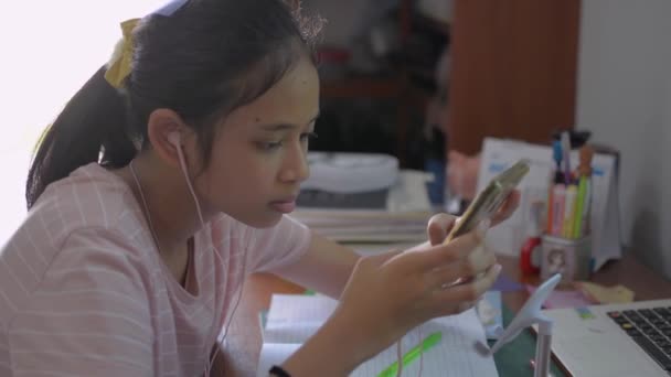 Roztomilý asijské dospívající student nosit sluchátka smskování zprávy na vzdálený učitel na smartphone. Střední škola dívka studium lekce on-line prostřednictvím mobilní aplikace během pandemické koronavirus situace. - Záběry, video