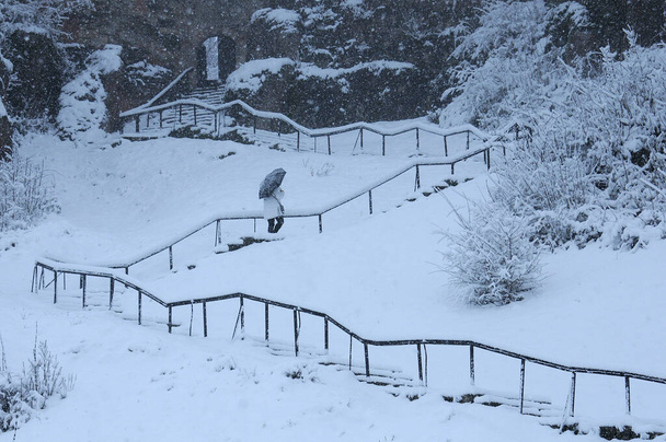 Άγνωστη γυναίκα με λευκό παλτό ανεβαίνει τα σκαλιά του φρουρίου κάτω από μια μαύρη ομπρέλα στη χιονοθύελλα. - Φωτογραφία, εικόνα