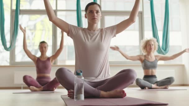 Full shot of young Caucasian woman wearing sportswear, sitting in lotus position on yoga mat in foreground of blurred people in fitness room, then closing eyes and joining palms together, meditating - Video
