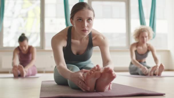 Eingesperrte junge kaukasische Frau in Sportkleidung sitzt mit geraden Beinen auf Yogamatte, streckt sich nach vorne, verschwommene Menschen üben im Hintergrund - Filmmaterial, Video