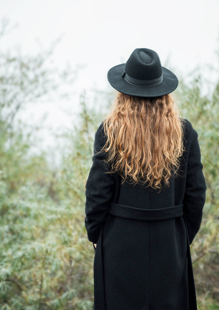 Portrét mladé atraktivní ženy v černém plášti a klobouku. Podzimní krajina, suchá tráva. Back vie - Fotografie, Obrázek