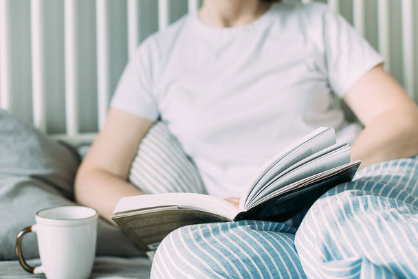Молодая брюнетка в белой футболке читает книгу и пьет кофе или чай на кровати дома. Концепция уютного, комфортного дома. Стиль жизни, подлинный момент - Фото, изображение