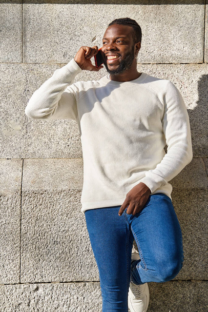 Счастливый молодой африканский мужчина, пользующийся мобильным телефоном и улыбающийся, прислонившись к стене. Латиноамериканцы разговаривают по телефону.  - Фото, изображение